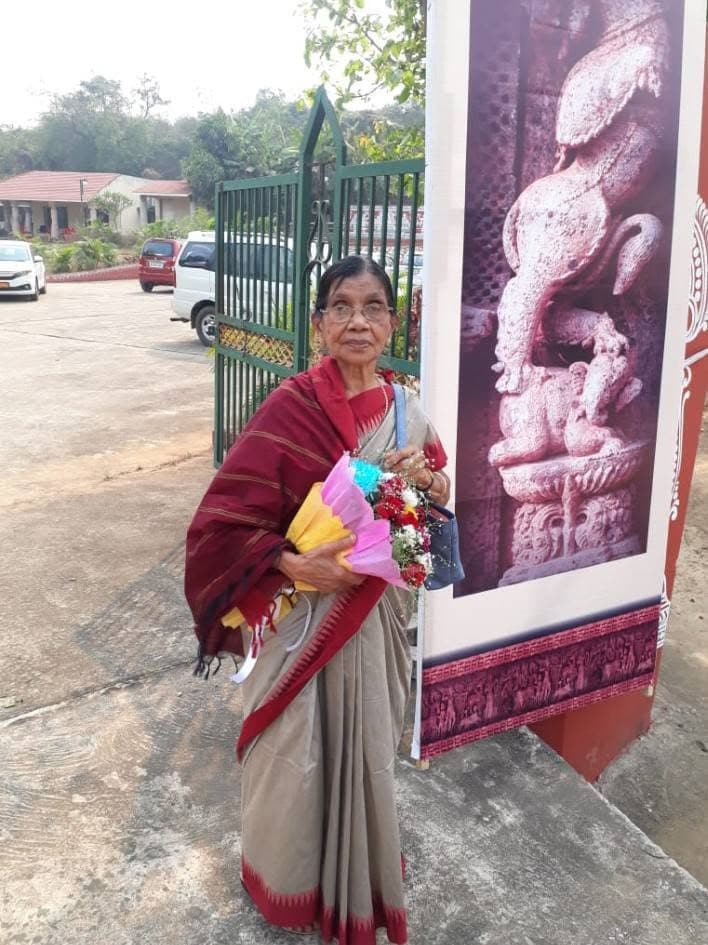 Basanta Kumari Samanta holding a bouquet of flowers beside a stone relief sculpture display at a heritage site