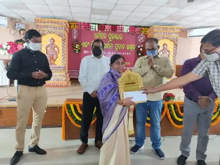 Basanta Kumari Samanta receiving a plaque on stage from a masked dignitary during a felicitation programme
